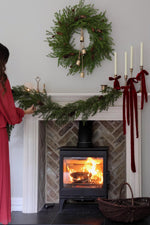 Person arranging an artificial Christmas wreath and garland on a fireplace mantel with festive decorations and long red ribbon. 
