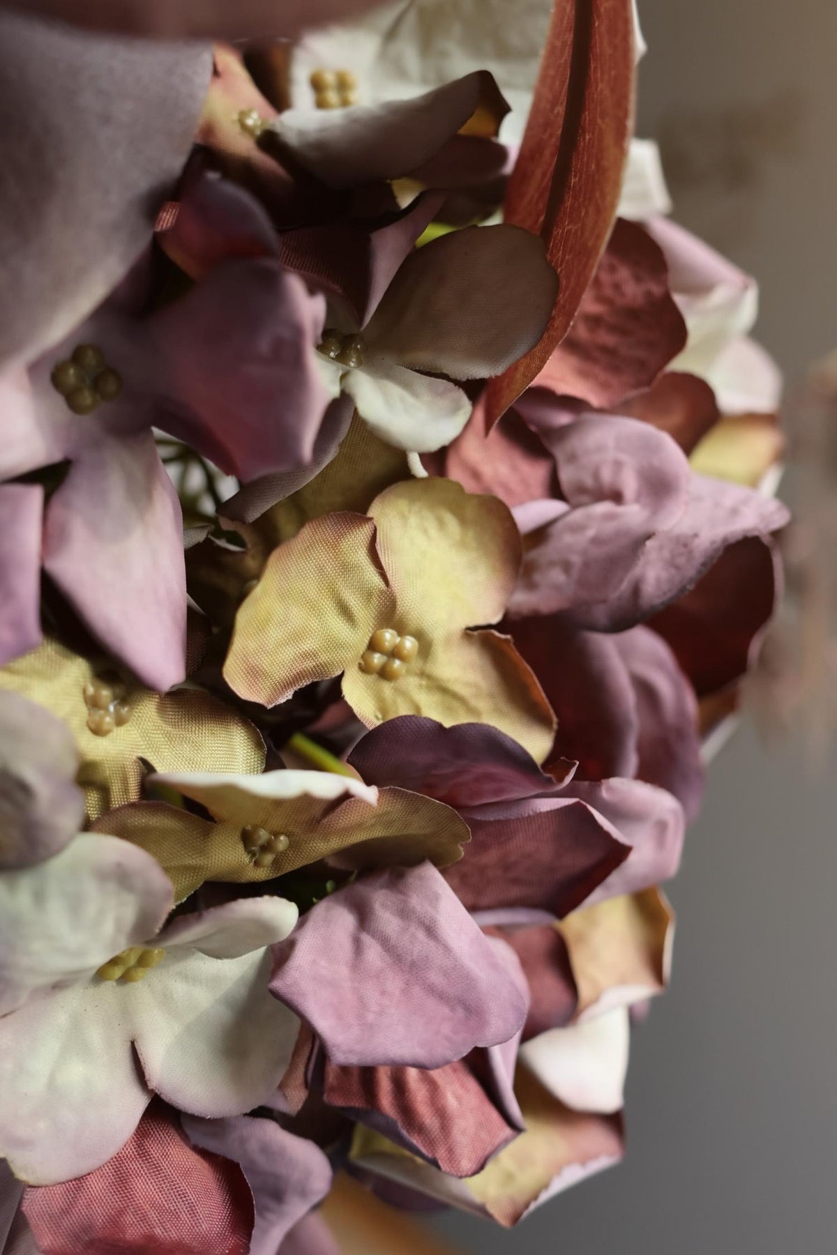 A close-up of an artificial dried look hydrangea in muted tones, including dusty pink, mauve, cream, and burgundy, with soft blurred background.
