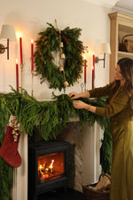 A cozy holiday fireplace scene with a green garland draped over a black wood-burning stove, a matching wreath on the mantel, red candles, a red Christmas stocking, and a basket of firewood on the floor.