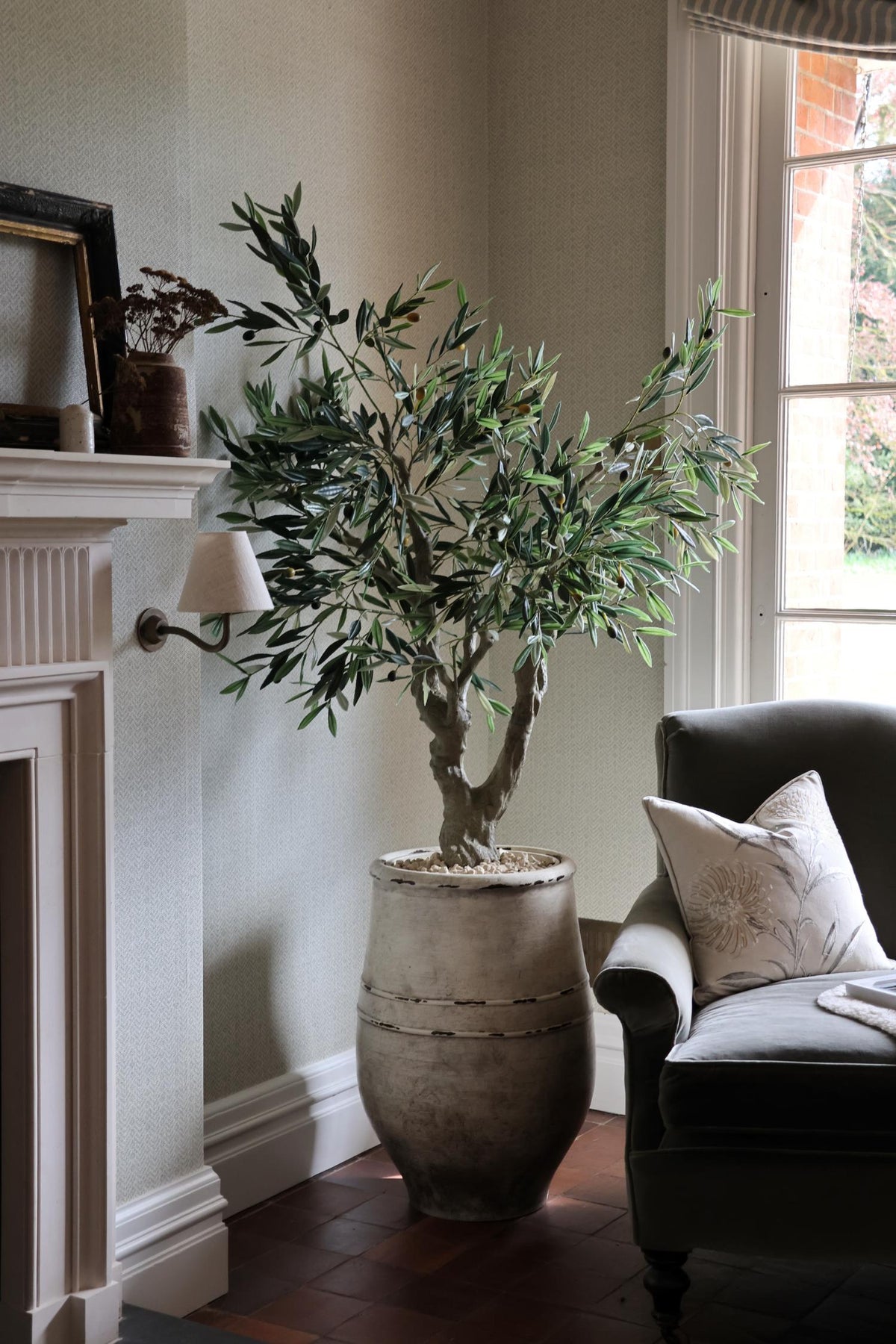 artificial potted olive tree in corner of a room in a tall rustic planter next to a fireplace and chair