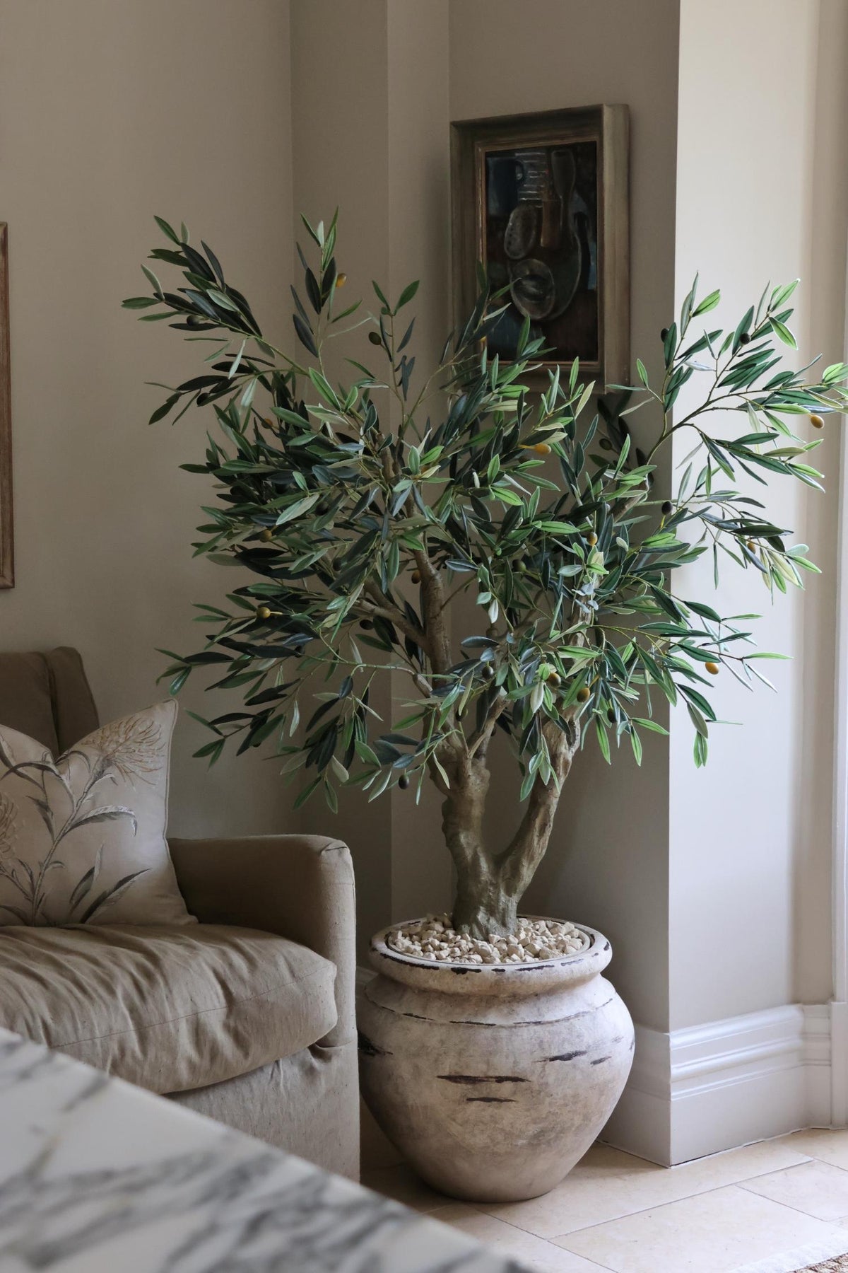 A faux olive tree with a realistic multi-stemmed trunk and lifelike leaves and olives, placed in a large pot with stones on top, situated in a room with a sofa and a framed picture on the wall.