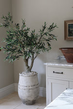 Potted artificial faux olive tree in a room with a dresser and decorative item on a shelf.