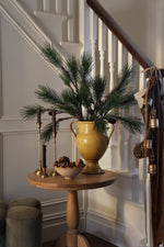 artificial pine branches styled in a yellow ceramic urn on a small round table in front of a white staircase 
