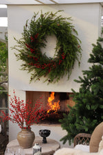 Large artificial red berry larix wreath above a lit open fire with a vase of red berries and christmas tree 