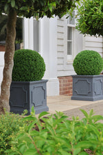 Two spherical premium artificial green topiary balls in gray planters in front of a white house.