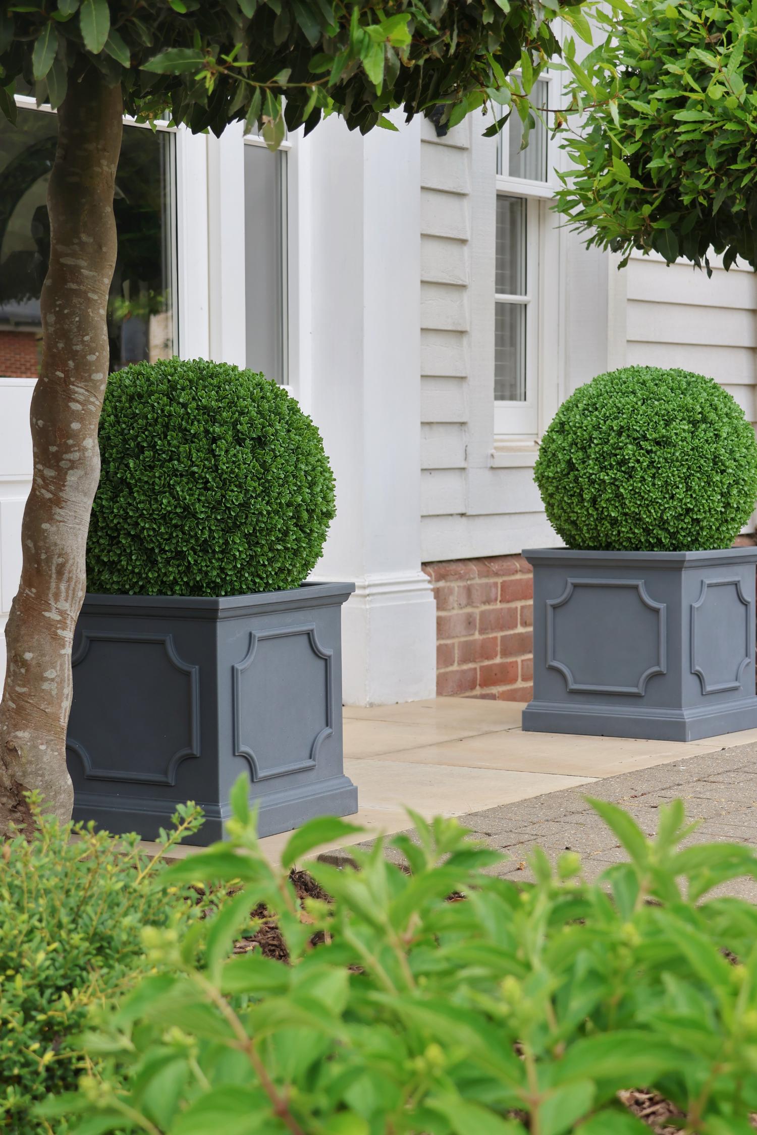 Two spherical premium artificial green topiary balls in gray planters in front of a white house.