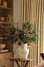 A rustic white ceramic vase filled with a natural pine branch arrangement featuring green needles, pinecones, and dried beige grasses, displayed on a wooden side table in a warm, neutral-toned room with beige curtains and wooden shelving.