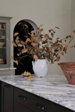 Artificial autumn foliage in a white vase on a marble countertop with a cabinet in the background.