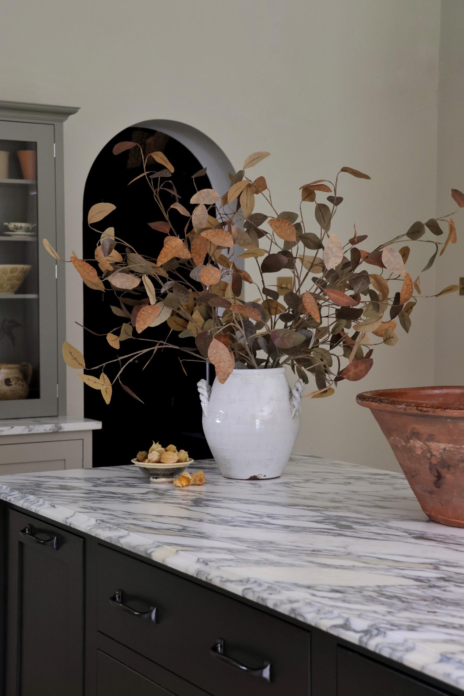 Artificial autumn foliage in a white vase on a marble countertop with a cabinet in the background.