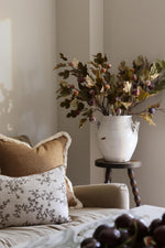 A rustic white ceramic vase filled with artificial autumn fig branches, displayed on a dark wooden side table next to a beige sofa with decorative pillows.