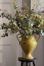 A yellow ceramic urn-style vase filled with dried green hydrangeas, eucalyptus, and other botanical stems, displayed on a small dark wooden stool in an indoor setting.