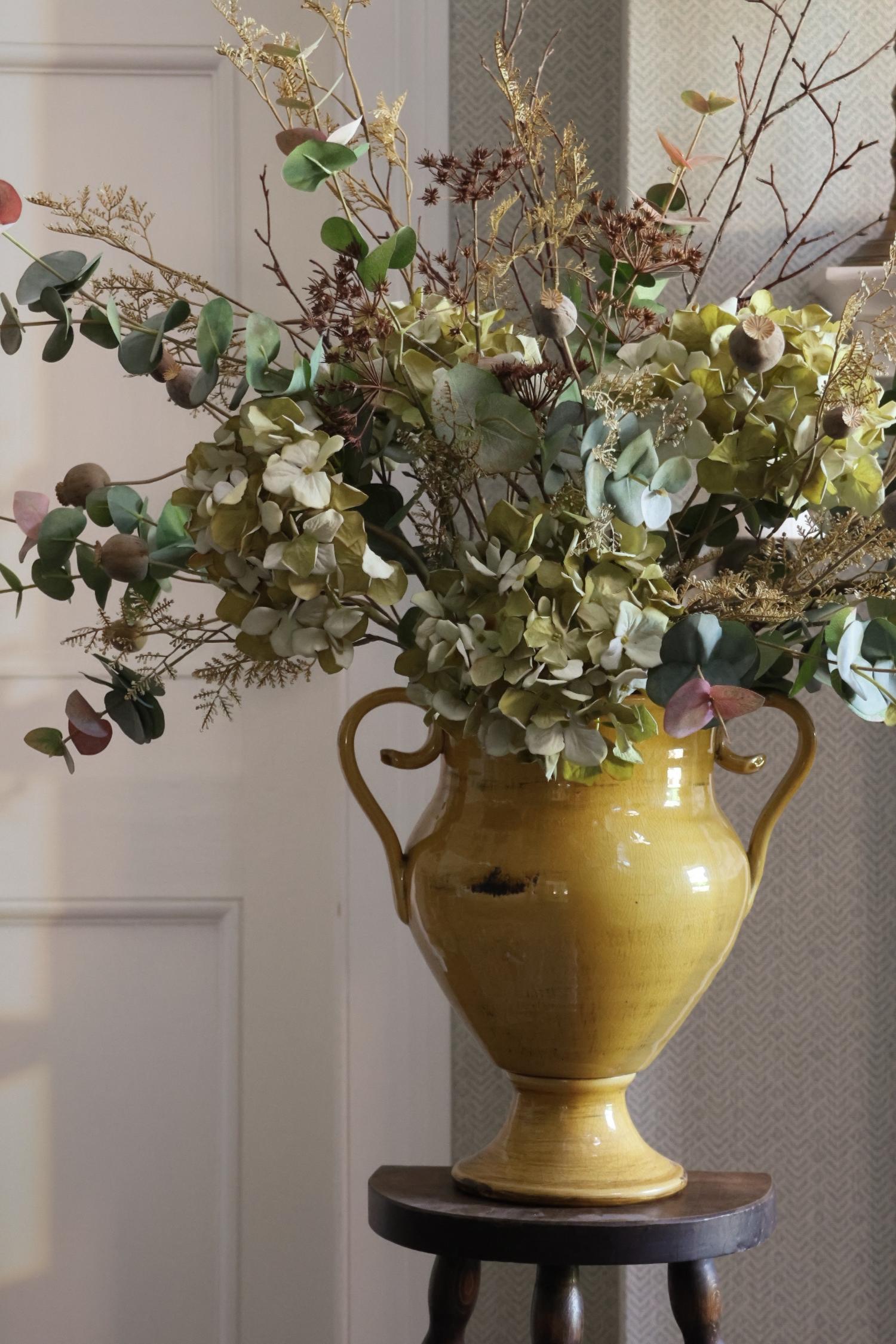 A yellow ceramic urn-style vase filled with dried green hydrangeas, eucalyptus, and other botanical stems, displayed on a small dark wooden stool in an indoor setting.