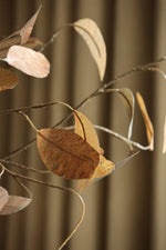 Artificial autumn foliage in large yellow urn with soft yellow curtains behind it 