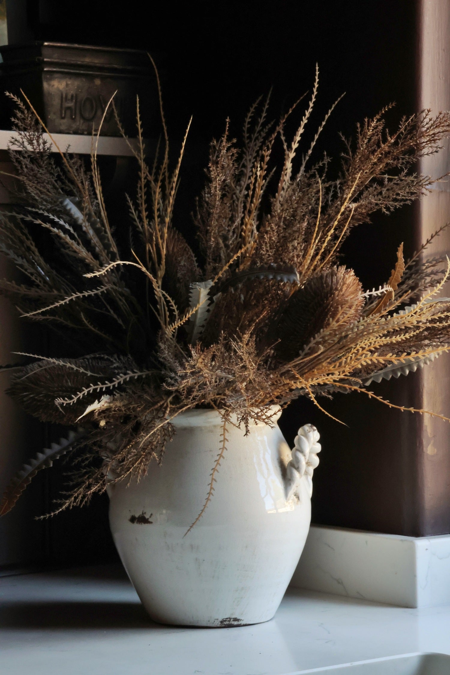A rustic white ceramic vase with a sculptural handle, filled with dried brown and tan botanical stems, set against a dark background.