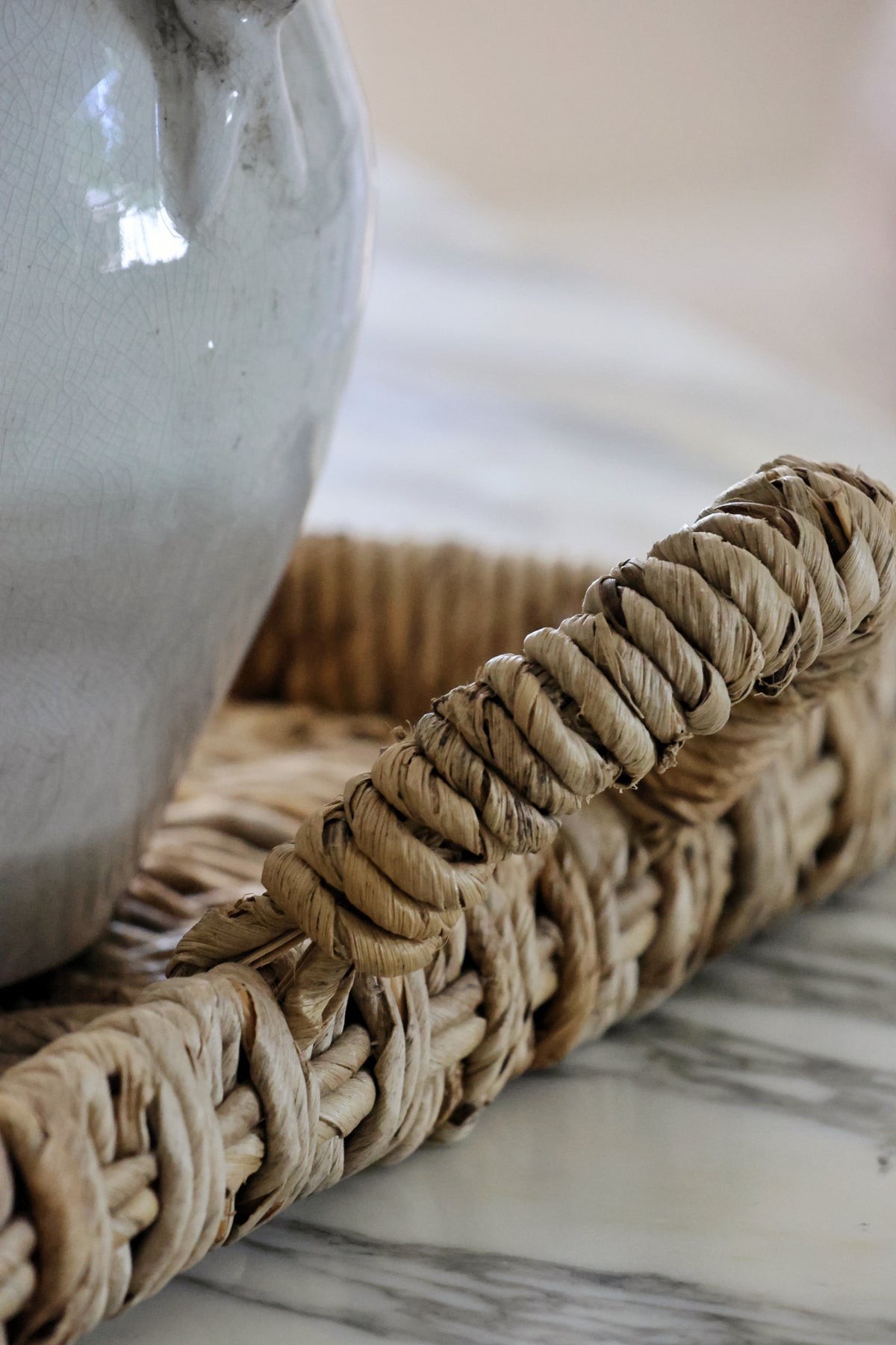 A close-up of a woven natural fiber tray with a thick twisted handle, placed on a light marble surface next to a white ceramic pitcher.
