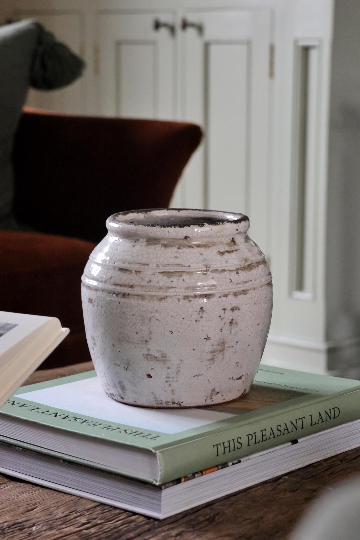 A rustic off-white ceramic pot placed on a book, with a blurred background featuring a cozy indoor setting.