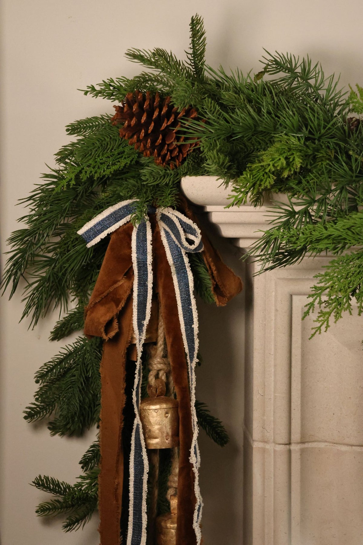 blue and brown decorative ribbon hanging in a bow with brass cowbells layered over an artficial christmas garland on the side of a limestone mantle
