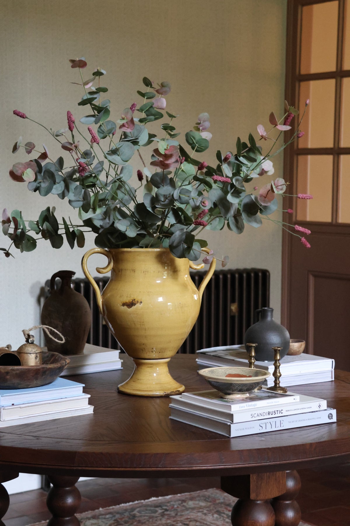 A yellow ceramic vase filled with artificial blush eucalyptus branches, styled on a dark wooden table with books and small decor objects in an indoor room setting.