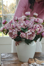 A person holding a vase with a faux blush peony stem and additional artificial flowers, set against a window backdrop.