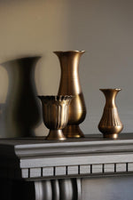 Three metallic gold decorative vases in different shapes displayed on a dark gray mantelpiece against a warm beige wall.