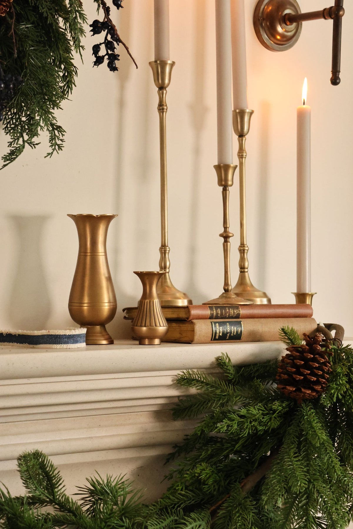 A styled holiday mantel scene featuring brass candlesticks, white taper candles, a small gold vase, books, pine garland, and a pinecone against a warm cream wall.