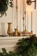 A styled holiday mantel scene featuring brass candlesticks, white taper candles, a small gold vase, books, pine garland, and a pinecone against a warm cream wall.