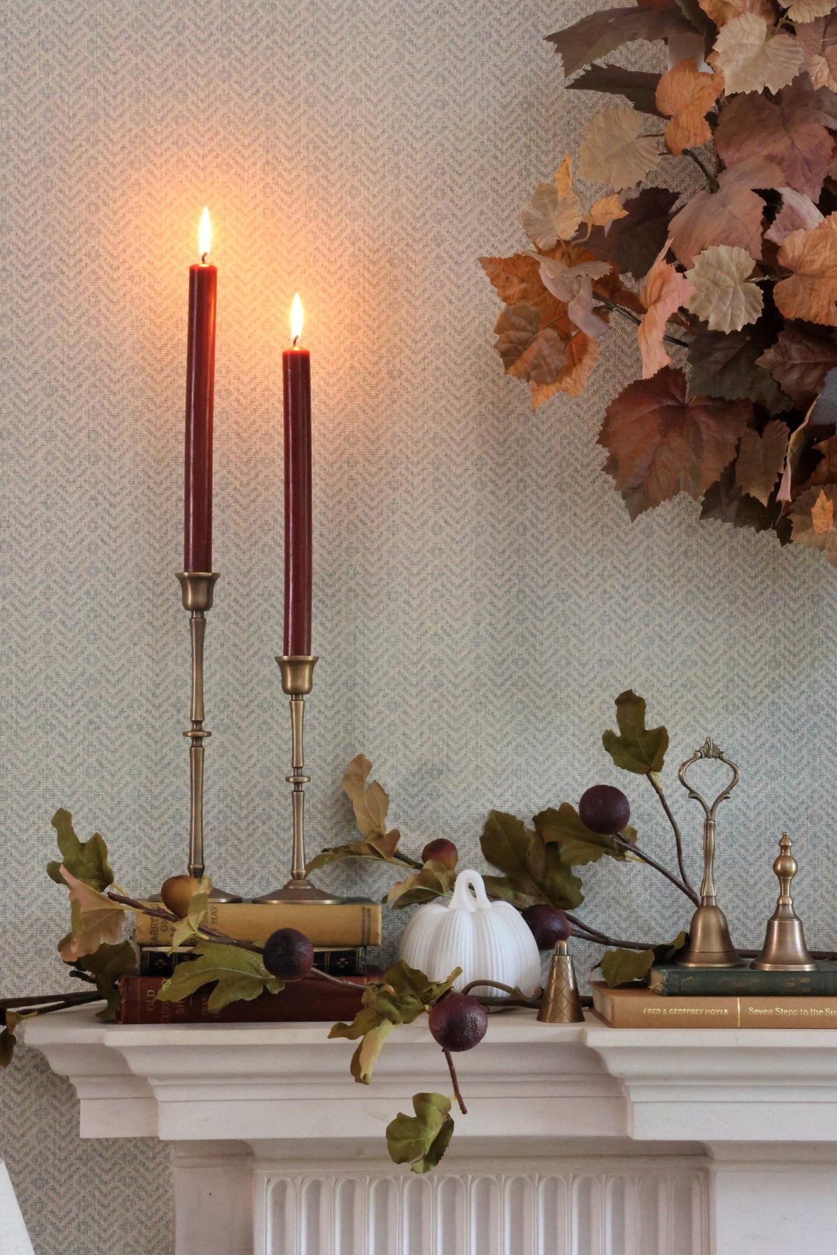 Decorative mantel with brass candle sticks, dark red lit candles, pumpkins, and fall leaves against a neutral wall.