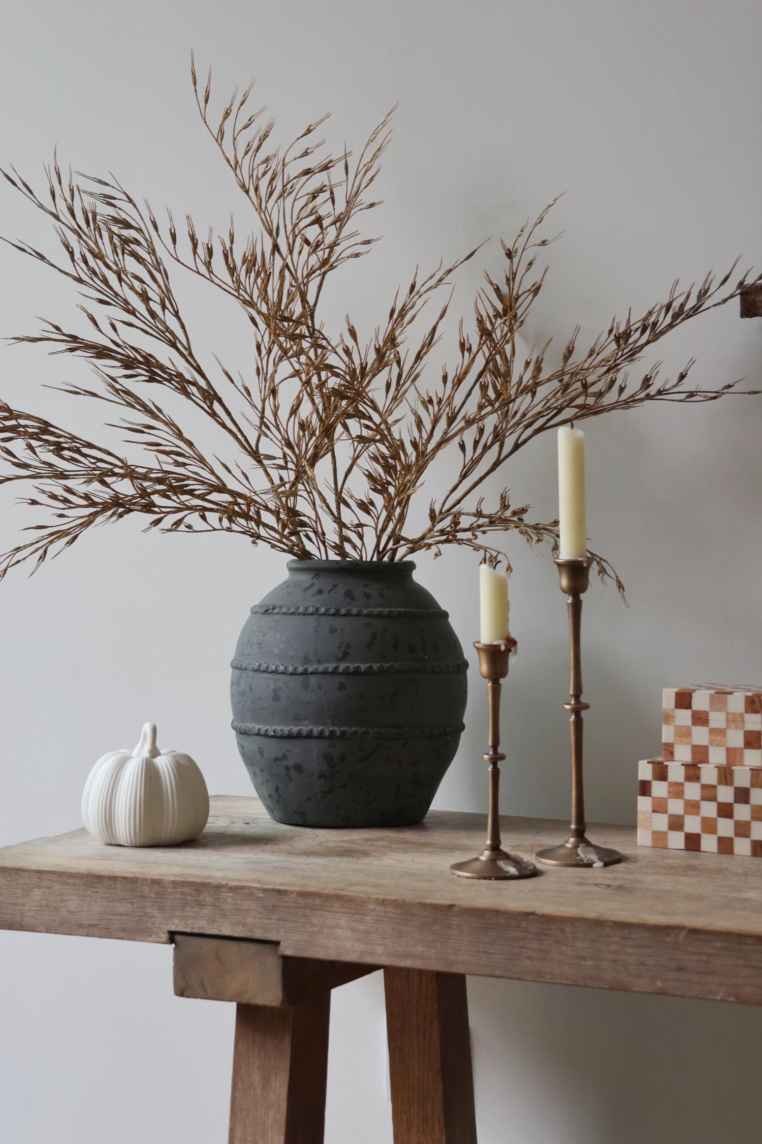 Decorative setup with a textured black vase, dried branches, candles, and a small pumpkin on a wooden surface.