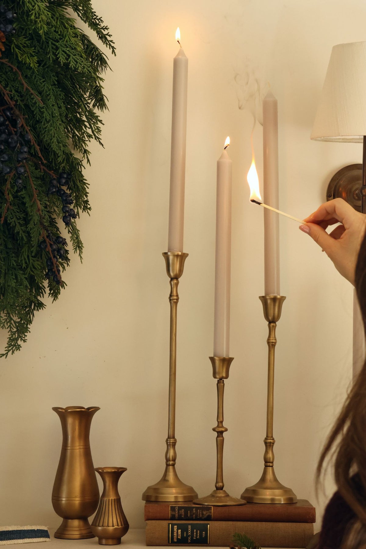 brass candlesticks sat ontop of some vintage books with a ladies hand and match about to light tall tapered candles 