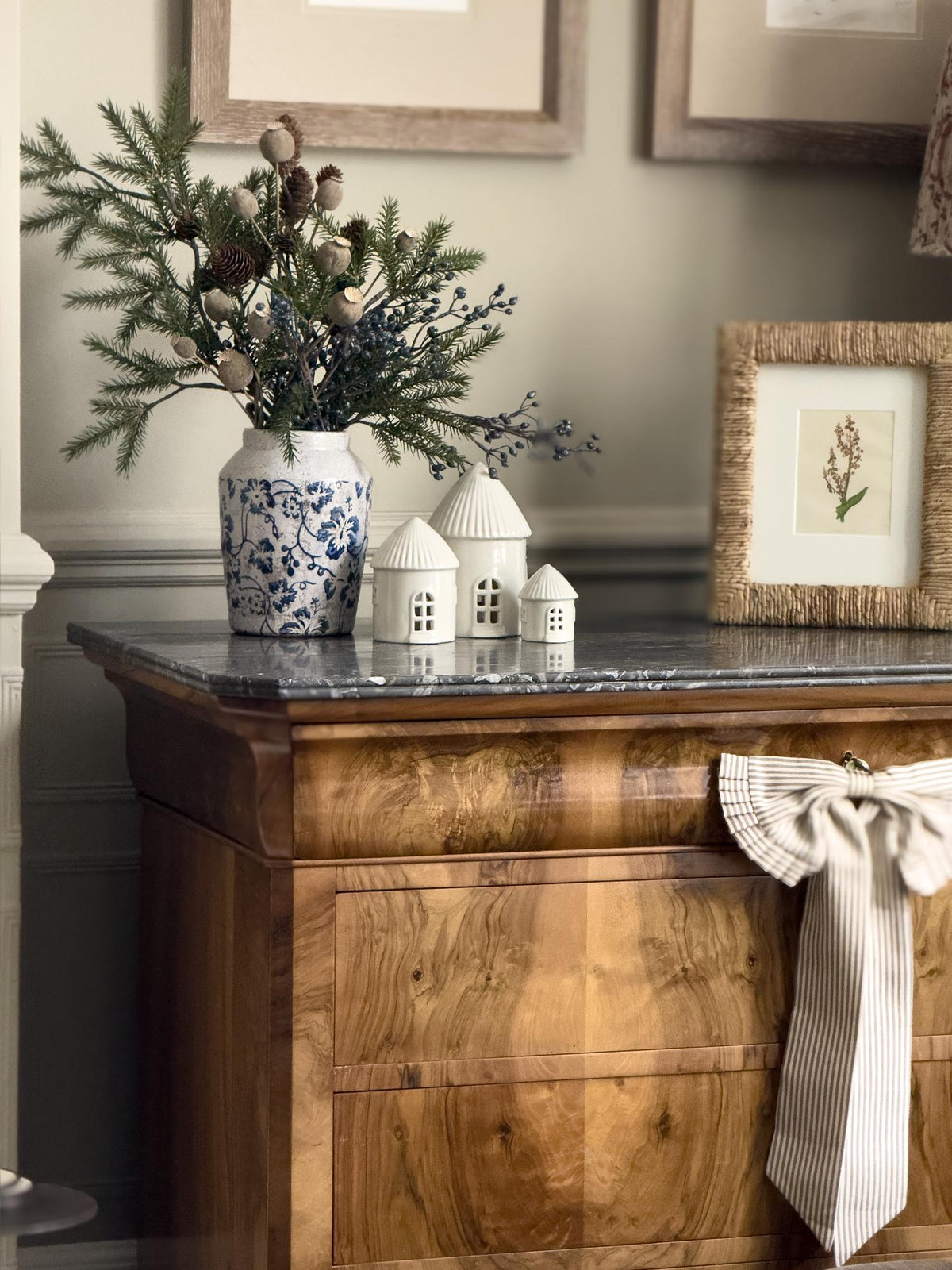 Decorative setting with a wooden cabinet, vase with greenery, small houses, and framed picture.