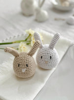 Two crochet bunnies, one beige and one white, with a gray nose and eyes, sitting on a white surface with a blurred background.