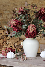 White vase with pink and green flowers on a wooden table against a stone wall.