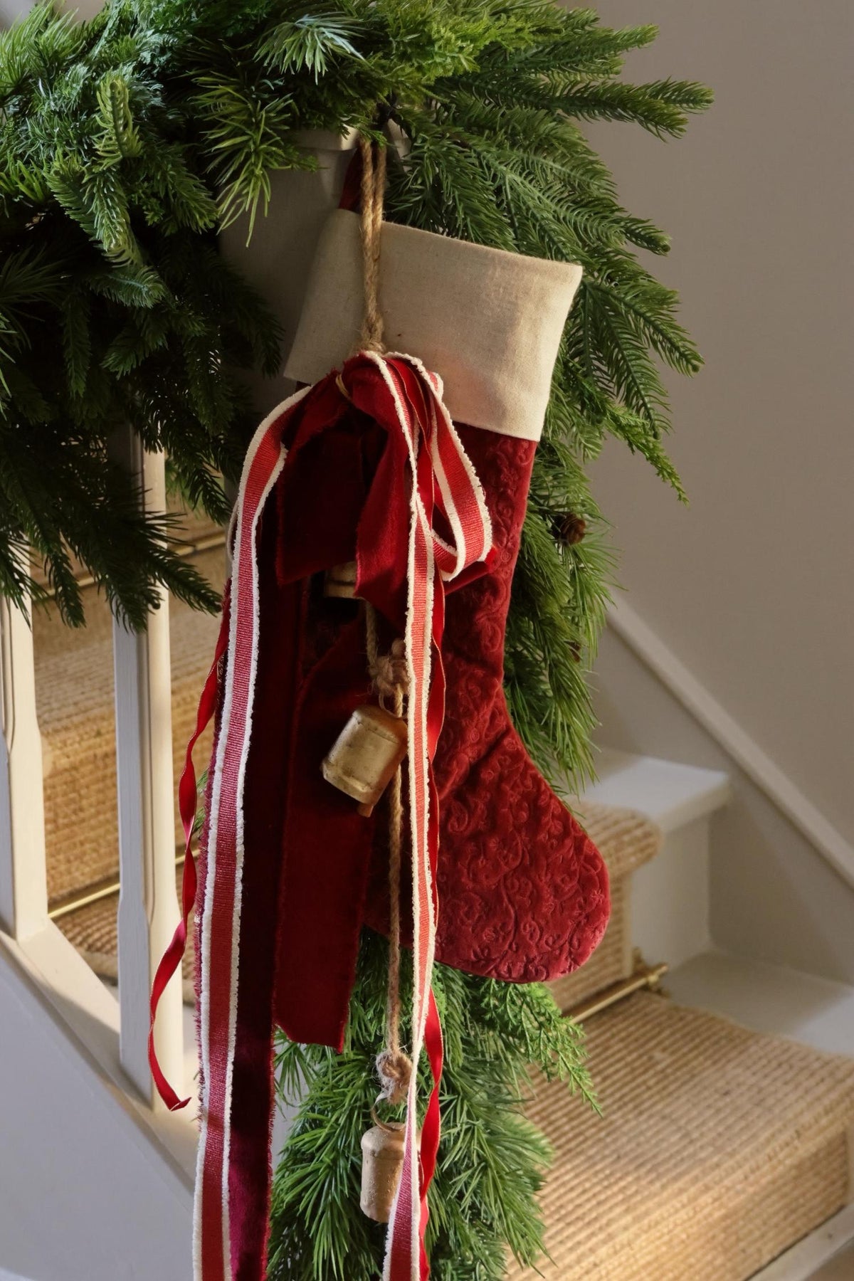 red velvet christmas stocking on staircase with green artificial garland and brass bells 