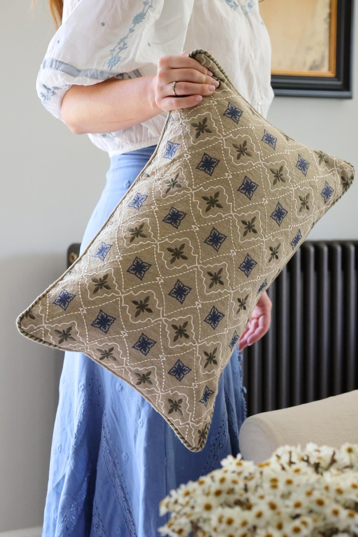 Person holding a patterned cushion in a room with a radiator and a plant.