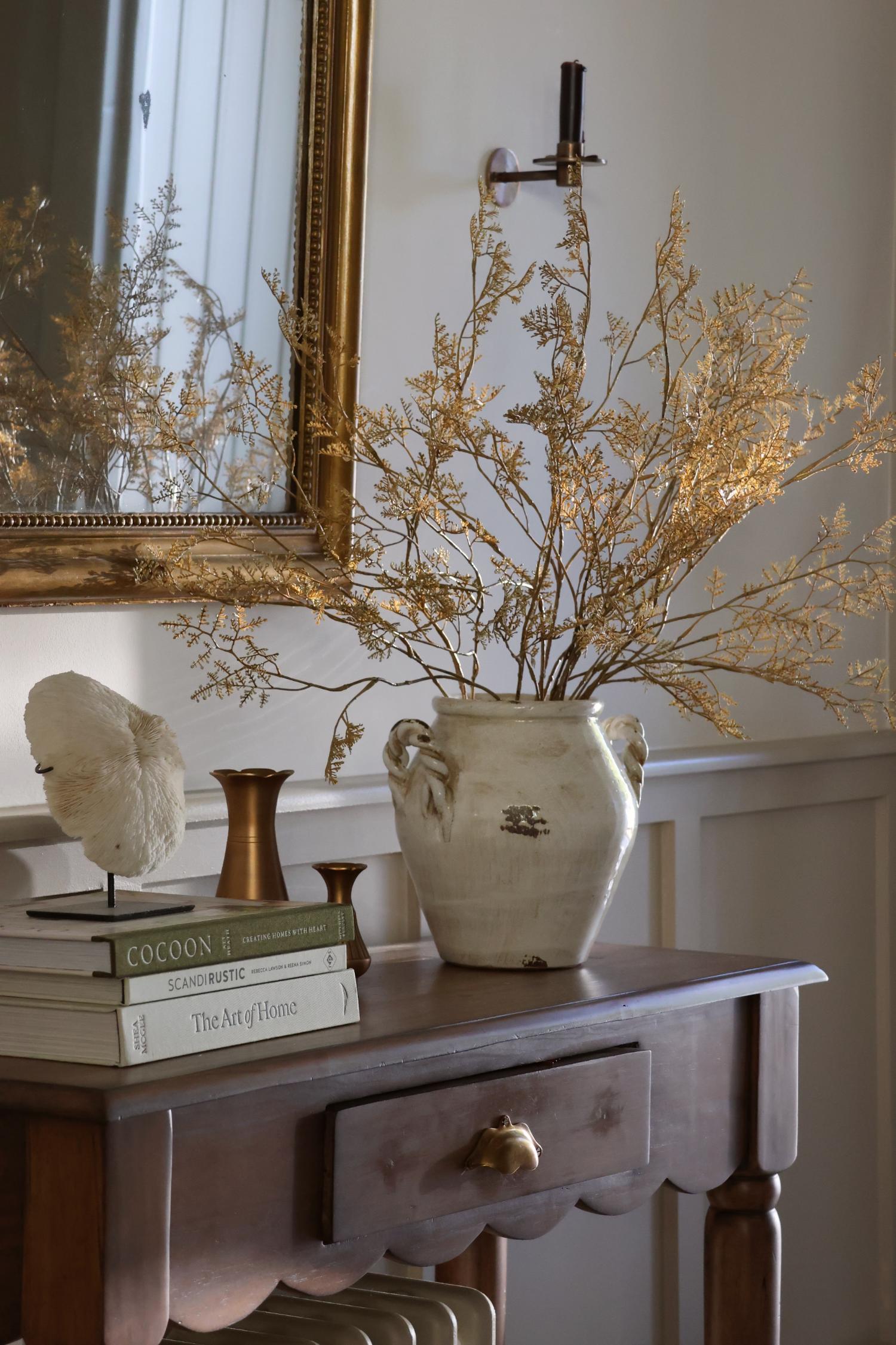 A rustic white ceramic vase filled with dried golden-beige branches, displayed on a dark wood console table in a styled interior setting with books and a mirror.