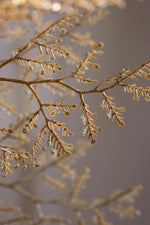 A close-up of delicate golden-brown branch-like decor with small icicles and clear hanging droplets against a soft blurred taupe background.