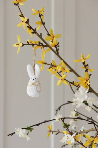 Decorative bunny ornament hanging from a branch with yellow and white flowers against a neutral background