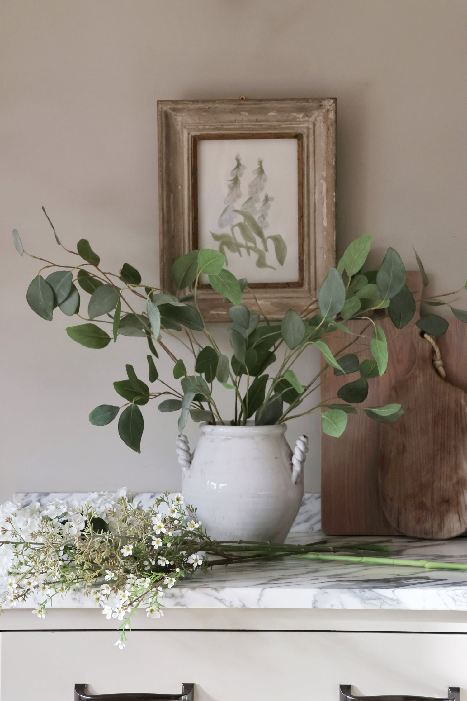 White vase with greenery on a marble surface with a framed picture in the background.