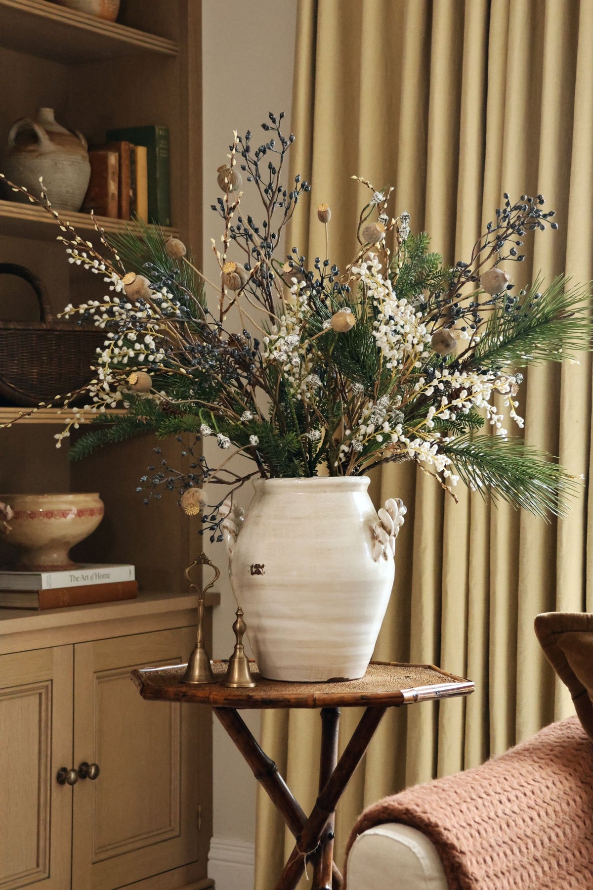a large faux blue berry arrangement with green foliage and white willow stems in a white ceramic rustic vase on a side table with beige curtains behind