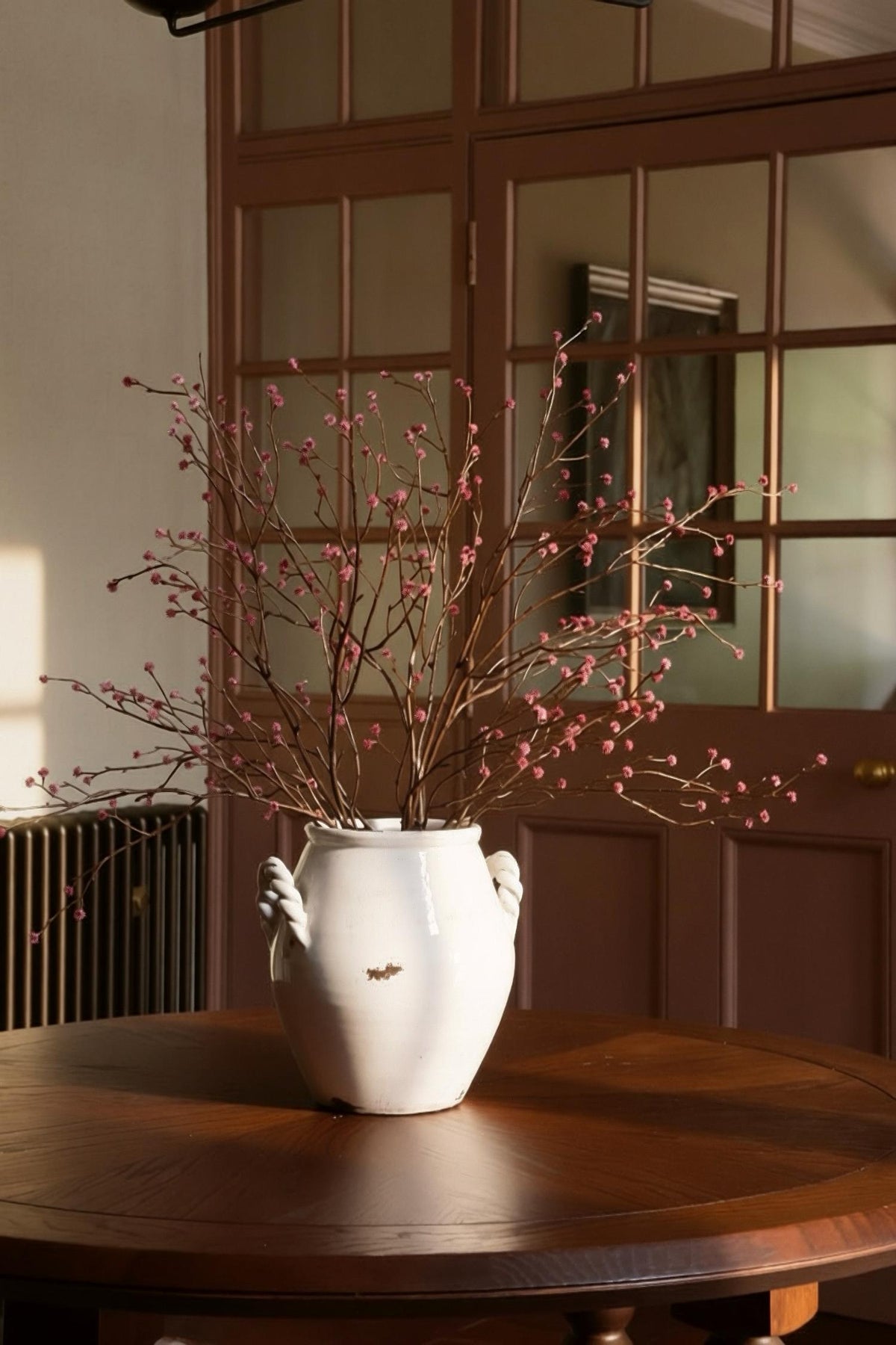A white ceramic vase with side handles, filled with artificial dried branches and small pink buds, placed on a round wooden table in front of a brown paneled door.