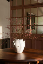 A white ceramic vase with side handles, filled with artificial dried branches and small pink buds, placed on a round wooden table in front of a brown paneled door.