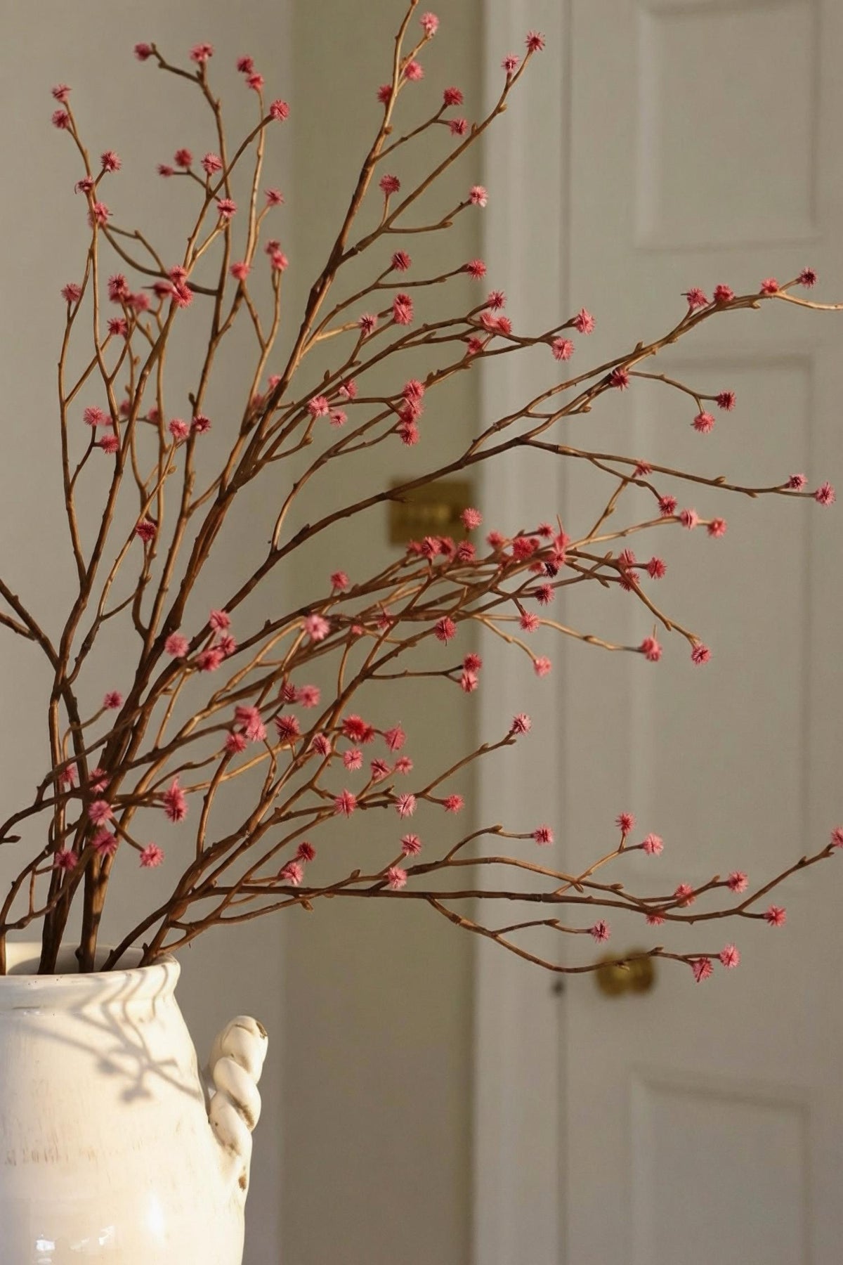 A white ceramic vase filled with faux dried brown branches and small pink buds, set against a softly blurred indoor background with a white door.