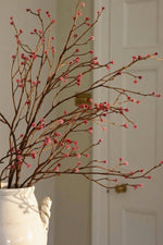 A white ceramic vase filled with faux dried brown branches and small pink buds, set against a softly blurred indoor background with a white door.