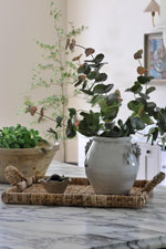 A rustic ceramic vase filled with artificial green eucalyptus branches, displayed on a woven tray over a marble countertop in a bright, modern interior setting.