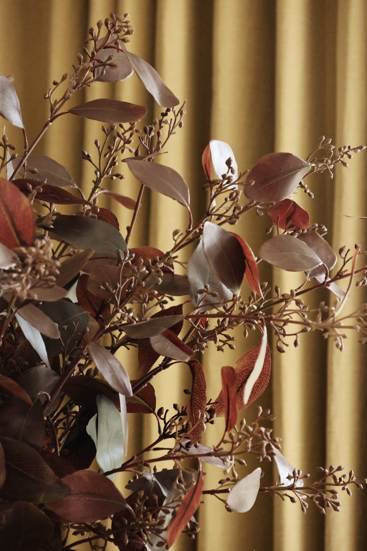 Artificial burgundy eucalyptus stems in a yellow urn vase with oak cabinetry and curtains behind 