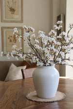 A faux cherry blossom stem in a white vase, placed on a wooden table with a round, beige placemat.