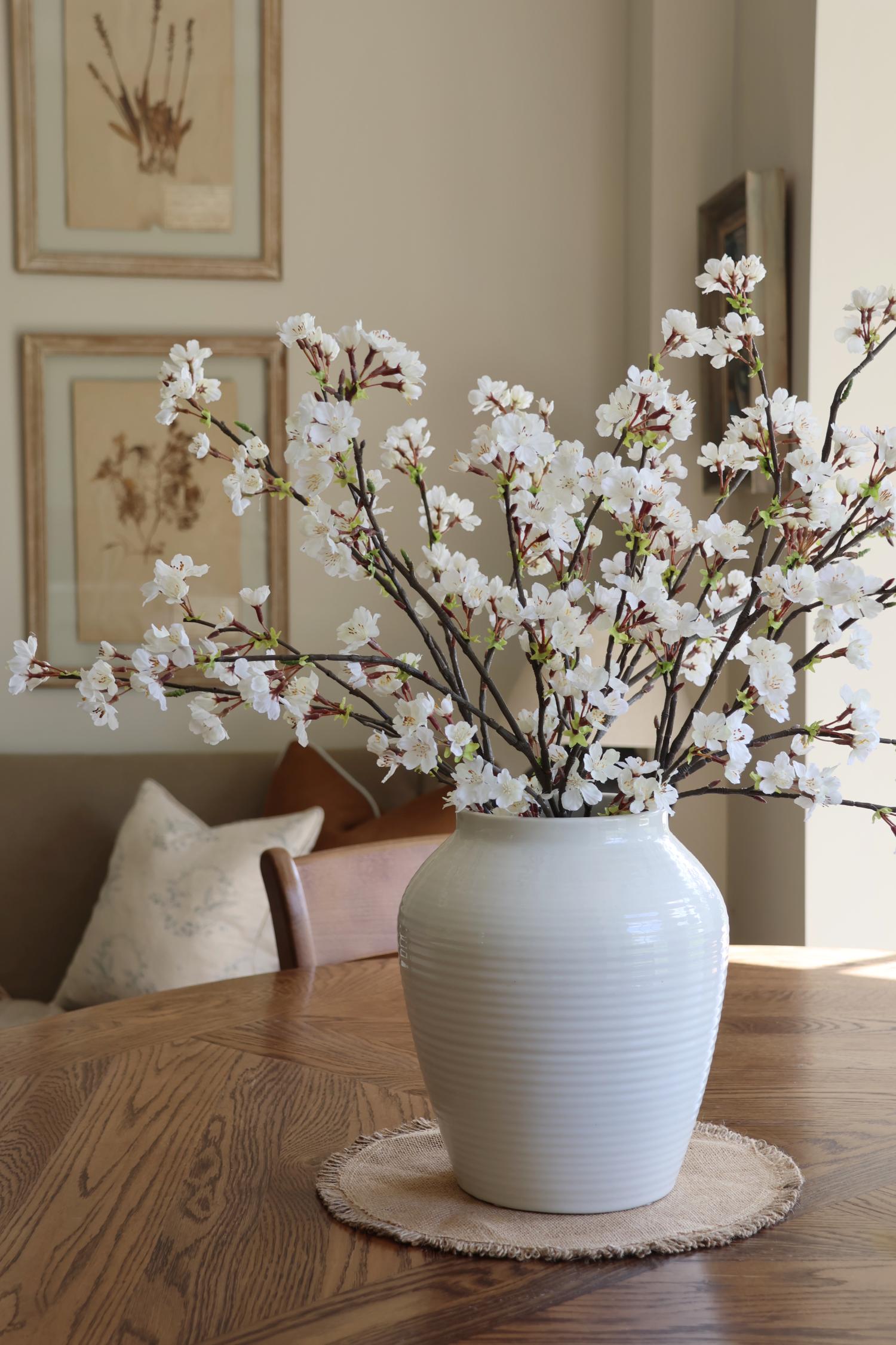A faux cherry blossom stem in a white vase, placed on a wooden table with a round, beige placemat.