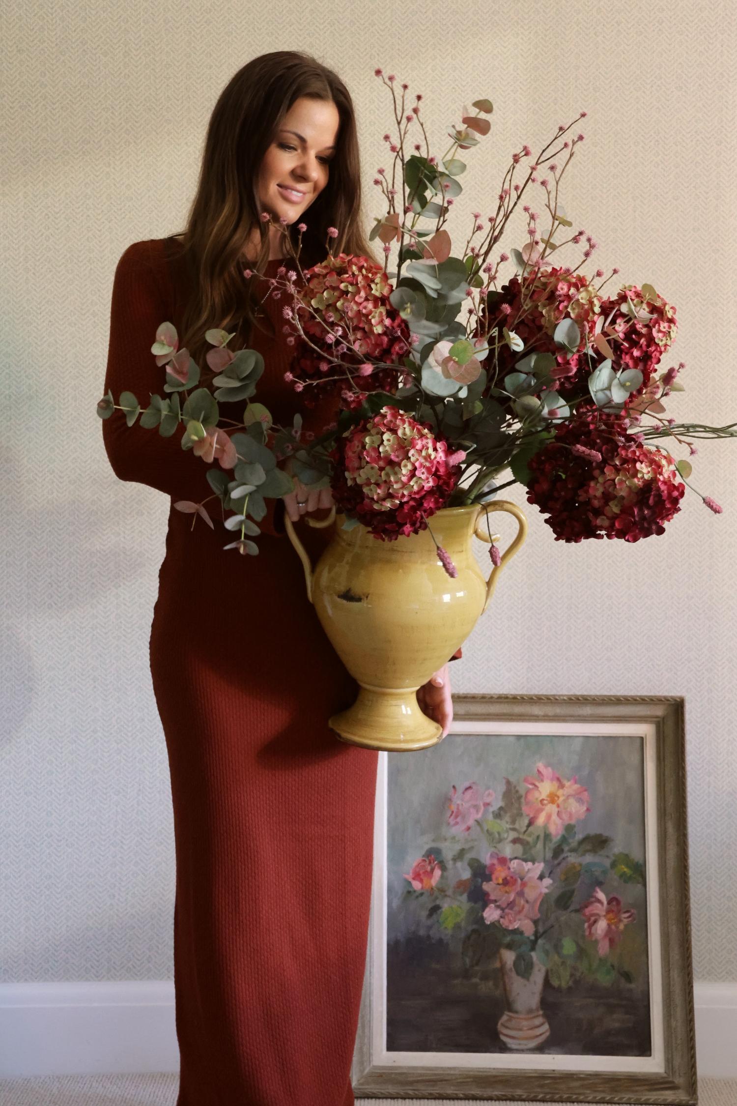 A woman in a long burgundy dress holds a large yellow ceramic vase filled with artificial deep red hydrangeas and eucalyptus stems, standing indoors next to a framed floral painting.