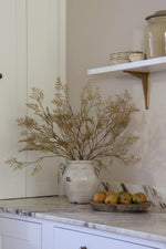 A rustic white ceramic vase filled with dried artificial beige branches on a white marble countertop, next to a wooden bowl of fruit, with a white wall and shelf in the background.