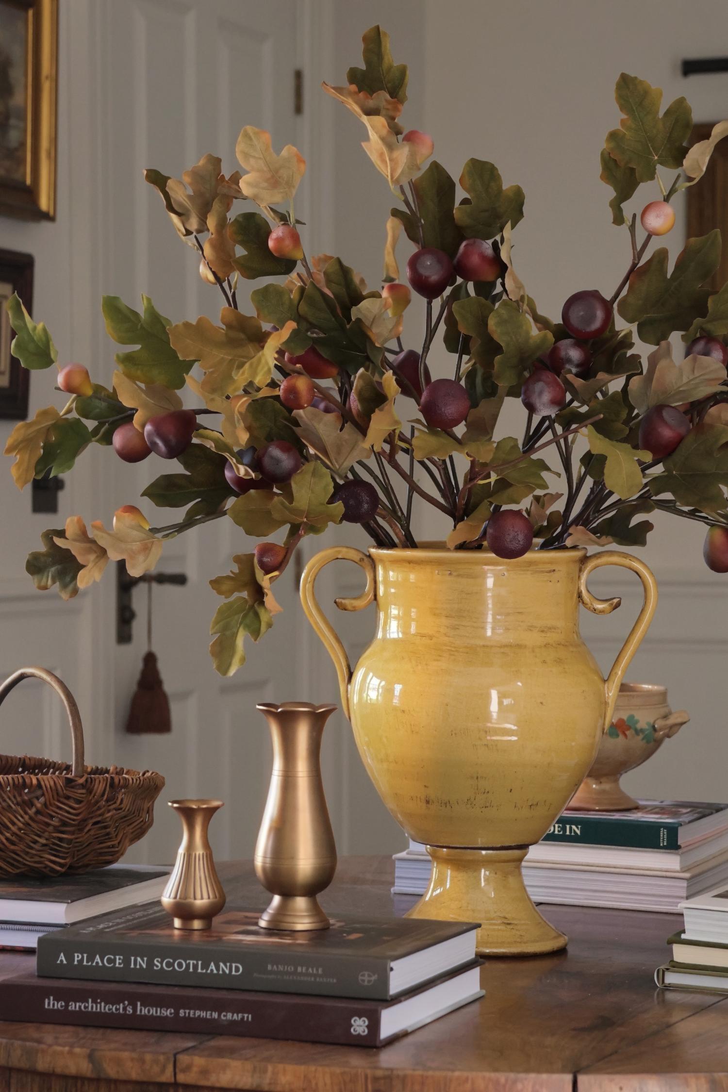 A large yellow ceramic urn-shaped vase filled with artificial autumn leaves and fig branches, displayed on a wooden table with books and small brass vases in an indoor setting.